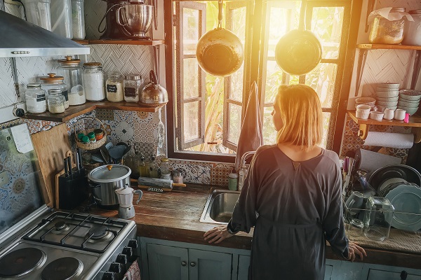 Mature woman standing next to a window in an old narrow cluttered kitchen with orange setting sun in a window. from behind, high angle.